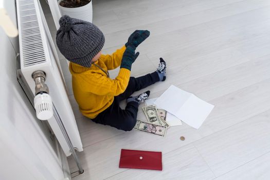 A little boy in a yellow sweater and hat is counting money and studying heating bills, near a heater with a thermostat..