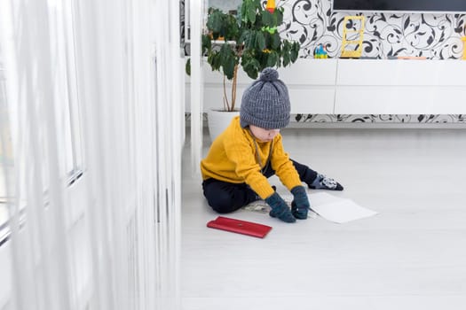 A little boy in a yellow sweater and hat is counting money and studying heating bills, near a heater with a thermostat..