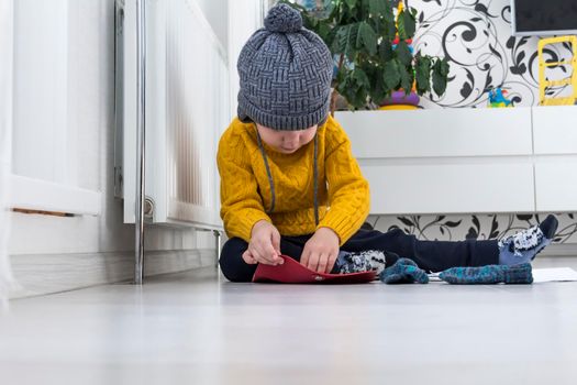 A little boy in a yellow sweater and hat is counting money and studying heating bills, near a heater with a thermostat..