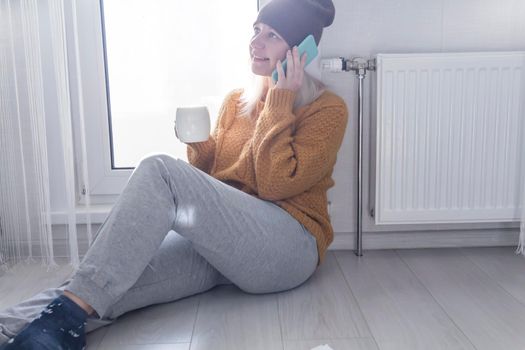 A young girl in a yellow sweater and hat is talking on the phone and drinking hot tea, near a heater with a thermostat..