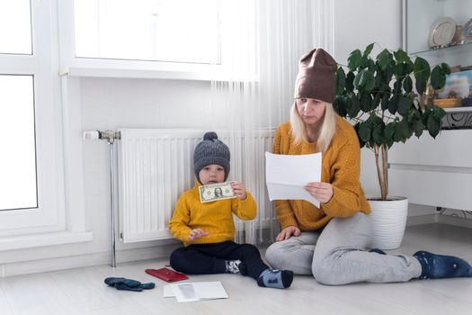 .A mother with a small child in a yellow sweater and hats is counting money and thinking how to pay bills and taxes, near a heater with a thermostat.