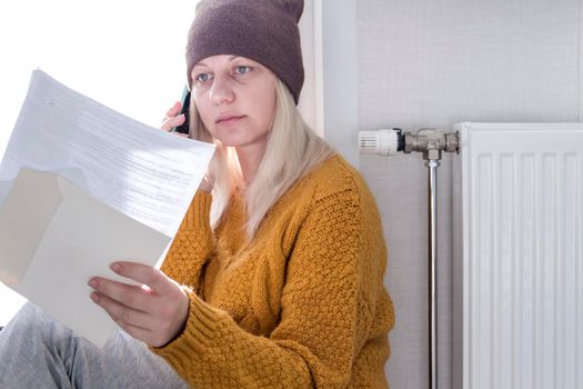 A young girl in a yellow sweater and a brown hat is sitting on the floor, counting money and thinking how to pay bills and taxes near a heater with a thermostat..