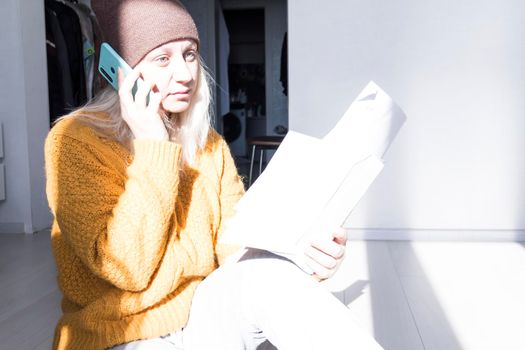 A young girl in a yellow sweater and hat is talking on a mobile phone and studying large bills and utility bills..