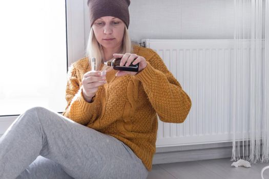 A young girl in a yellow sweater drinks medicine and sits on the floor near the radiator..