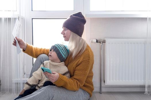 .A mother with a small child in a yellow sweater and hats is counting money and thinking how to pay bills and taxes, near a heater with a thermostat.