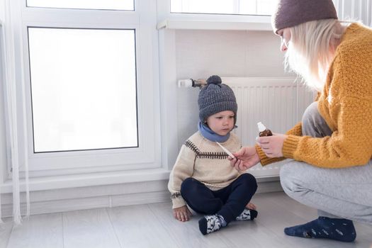 A young mother in a yellow sweater measures the temperature of the child and gives medicine for the disease..