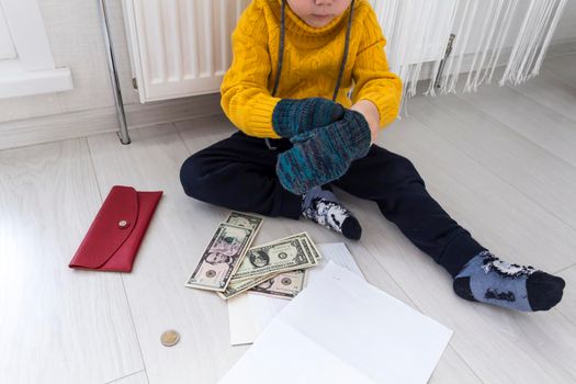 A little boy in a yellow sweater and hat is counting money and studying heating bills, near a heater with a thermostat..