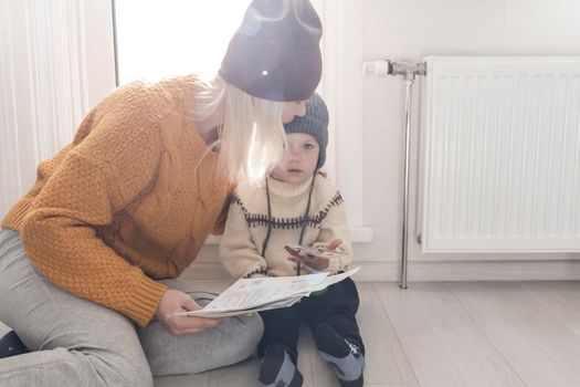.A mother with a small child in a yellow sweater and hats is counting money and thinking how to pay bills and taxes, near a heater with a thermostat.