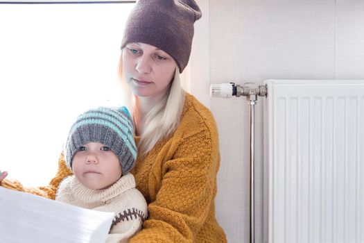 .A mother with a small child in a yellow sweater and hats is counting money and thinking how to pay bills and taxes, near a heater with a thermostat.