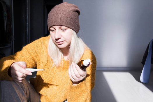 A young girl in a yellow sweater and a brown hat holds a thermometer in her hand and a jar of medicine..