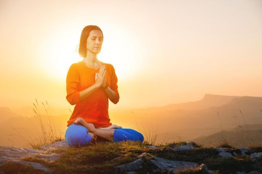 Yogini in the lotus position in full face sits on a cliff against the backdrop of the sunset sky with mountains, prays.