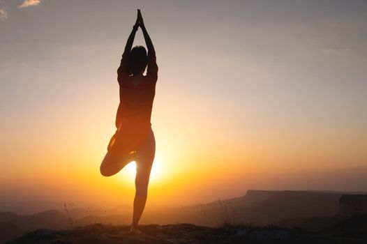 Silhouette of a young woman in a tree pose outdoors in the mountains watching the sunset, yoga balance near the cliff.