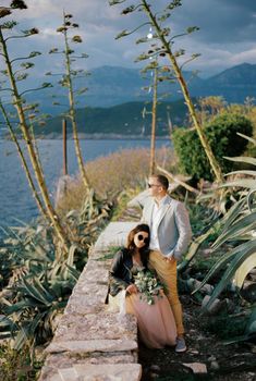 Bride sitting on a stone fence leaned against the standing groom. High quality photo