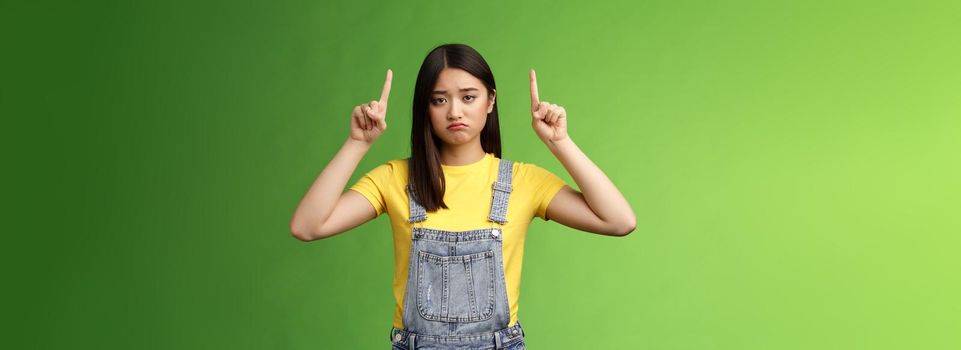 Pity cute sad asian girl brunette, grimacing pulling sorrow upset face frowning bothered, pointing fingers up regret awful situation, indicate top promo, stand green background unhappy.