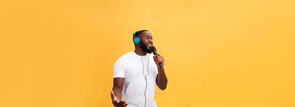 Portrait of cheerful positive chic. handsome african man holding microphone and having headphones on head listening music singing song enjoying weekend vacation isolated on yellow background.