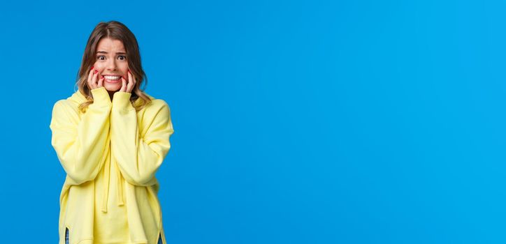 Embarrassed and afraid young timid blond caucasian female in yellow hoodie panicking, biting fingernails from fear and worries, looking scared with alarmed expression, blue background.