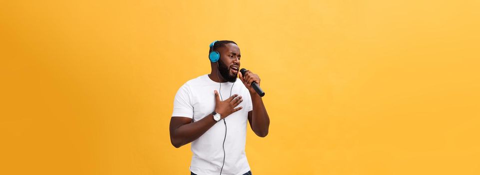 Portrait of cheerful positive chic. handsome african man holding microphone and having headphones on head listening music singing song enjoying weekend vacation isolated on yellow background.