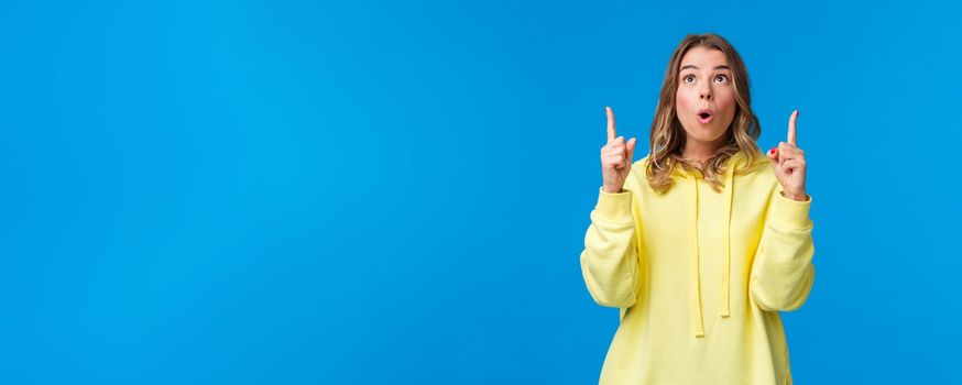 Dreamy and impressed startled cute caucasian female in yellow hoodie, make surprised and curious face with lips folded in wow, look and pointing up, top advertisement, blue background.