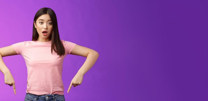 Shocked upset cute asian surprised girl, drop jaw shook disappointed, gasping disappointed pointing down amazed, indicating bottom advertisement, judging, negative opinion, purple background.