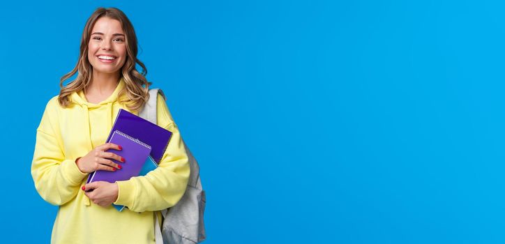Cheerful pretty blond girl smiling at camera, carry backpack and notebooks, papers for studying, learning new language at courses, standing joyful over blue background. Education and people concept