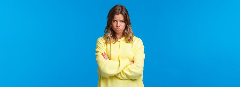 Gloomy and moody cute blond teenage girl look from under forehead and sulking at camera, feeling offended or upset, cross hands chest insulted waiting apology, blue background.