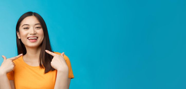 Close-up proud boastful cute asian girl telling story pointing herself, smiling broadly, bragging new haircut, standing pleased, delighted tell big news, stand blue background upbeat.