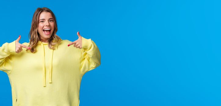 Confident, friendly-looking smiling blond girl in yellow hoodie pointing at herself, suggest own help, want volunteer or particiapte, say pick me, standing blue background.