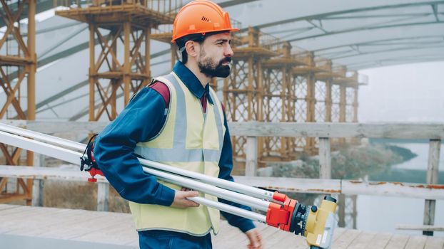 Side view of geodesy specialist walking in building area holding equipment wearing uniform on workday. Professionals and workplace concept.