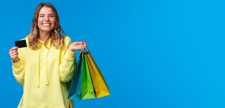 Happy smiling pretty girl using her credit card, deposit money for shopping, holding bags with clothes and grinning delighted, finally getting ready for summer vacation, blue background.