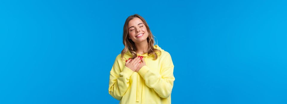 Romantic alluring blond caucasian girlfriend in yellow hoodie, press hands to heart and close eyes with lovely smile recall beautiful memory, imaging perfect date, standing blue background.