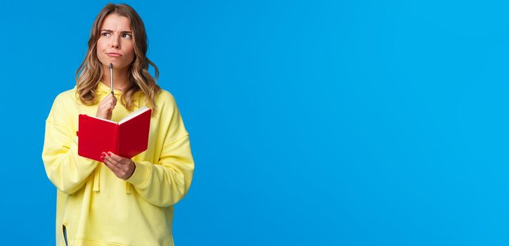 Waist-up portrait of cute european blond female in yellow hoodie making plans, solving homework or write diary, touching lip with pen pondering look upper left corner thoughtful, hold red notebook.