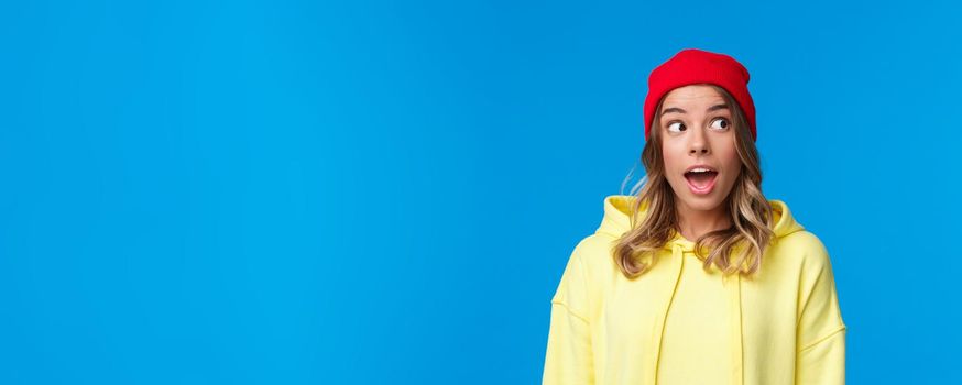 Excited good-looking caucasian female student in red beanie and yellow hoodie, look upper right corner and open mouth fascinated, hear interesting gossip, standing blue background.