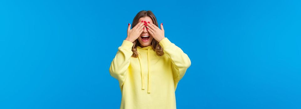 Girl waiting for friends tell her open eyes as shut it with hands and count ten during b-day party, girlfriend bring surprise gift, smiling with excitement, playing peekaboo, stand blue background.