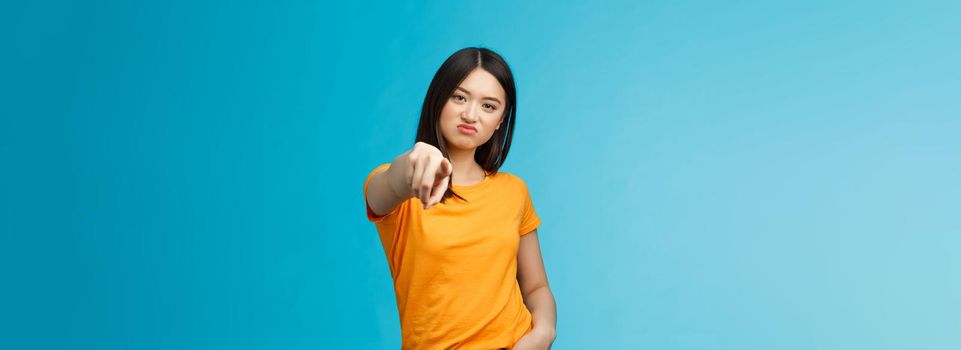 Disappointed attractive asian girl frowning, grimasing upset, pointing index finger camera upset unsatisfied, disliking you, accuse someone, blame person, standing displeased blue background.