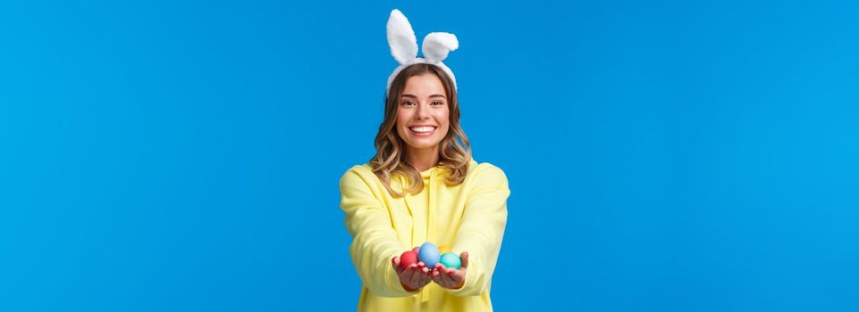 Holidays, people and fun concept. Charismatic cute blond girl holding coloured Easter eggs, wearing bunny ears and smiling joyfully, congratulate family with holy day, blue background.
