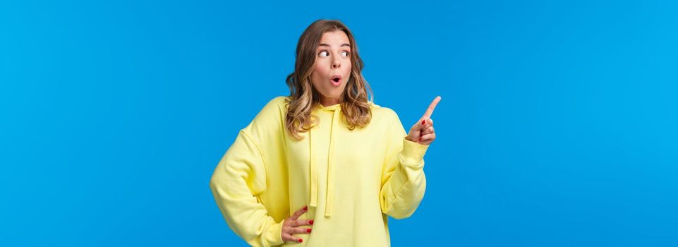Thrilled speechless good-looking blonde female in yellow hoodie react to something shocking and interesting, pointing finger and looking left saying wow, gossiping, stand blue background.