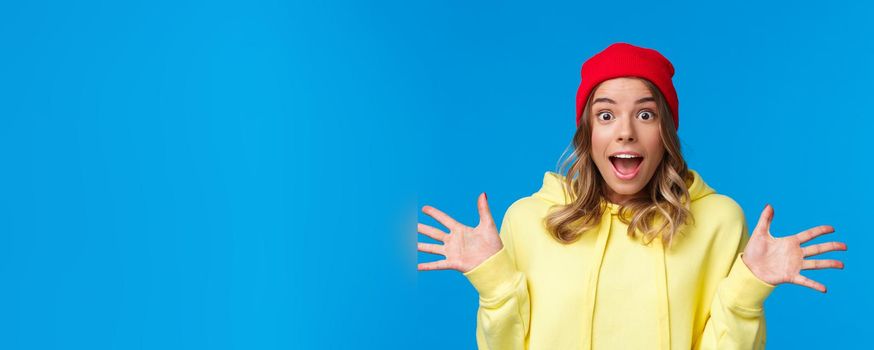 Close-up portrait thrilled and joyful amused european female in red beanie and yellow hoodie, listening to wonderful awesome news, spread hands sideways excited, stand blue background.