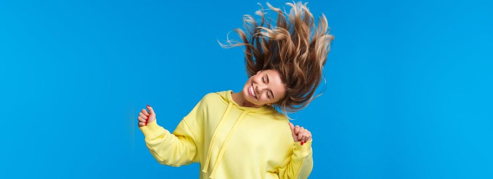 Carefree relaxed and joyful beautiful blond girl shaking head, dancing and jumping from happiness, having fun at awesome party or music concert, standing blue background.