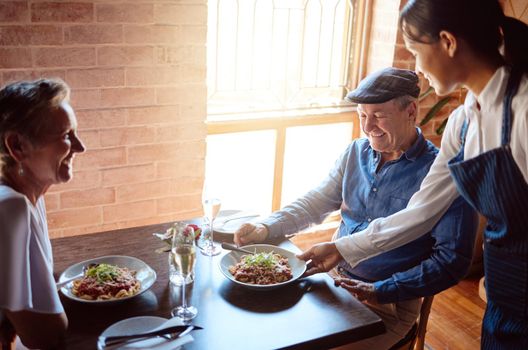 Happy senior couple at restaurant, waitress service with smile, and ready to eat healthy food with glass of champagne to celebrate anniversary. Woman in hospitality, serve lunch and drinks to people.