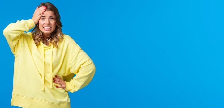 Embarrassed and worried girl bumped into car, feel anxious and concerned, touch head troubled, grimacing with awkward grin, face problematic situation, stand blue background.