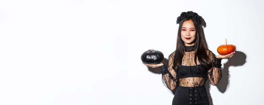 Pleased smiling asian woman celebrating halloween, wearing witch costume, holding pumpkins, standing over white background.