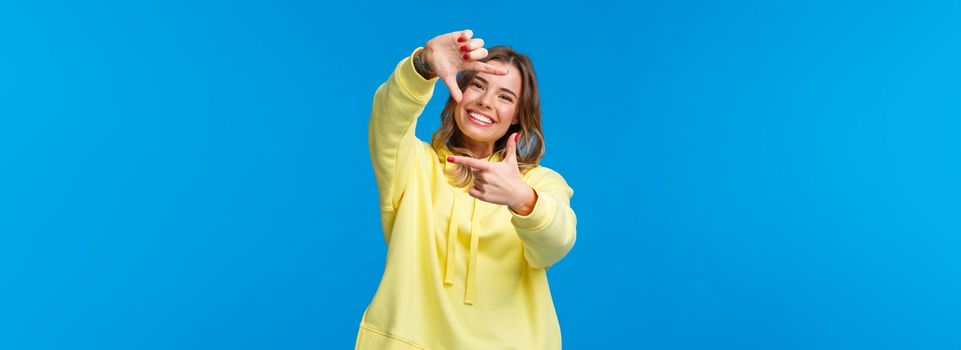 Inspiration, lifestyle and people concept. Portrait of cheerful creative blond girl searching perfect angle for shot, look at camera through frame gesture and smiling, blue background.