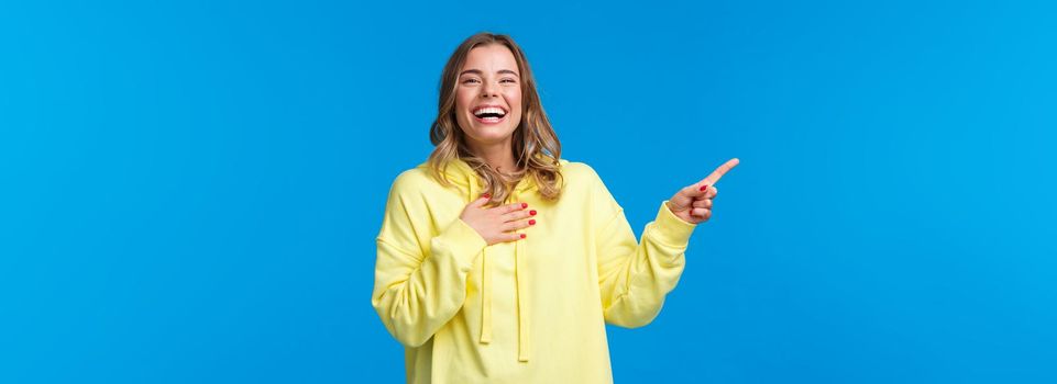 Happy and amused good-looking blond female in yellow hoodie, laughing out loud over something hilarious on upper left corner of blue background, smiling and looking entertained camera.