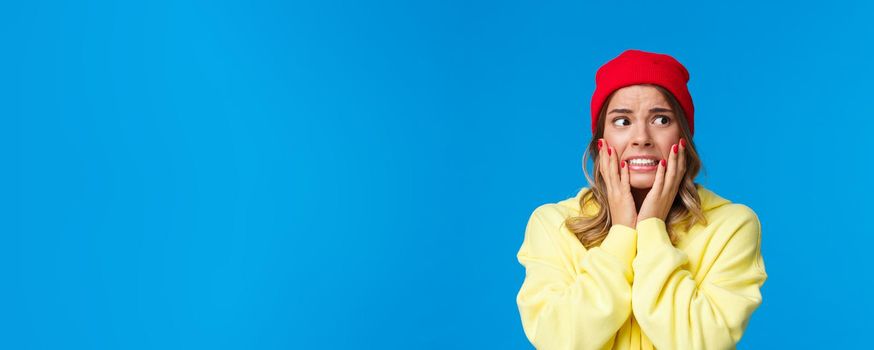 What have I done. Worried and concerned anxious caucasian female in red beanie and yellow hoodie, hold hands on cheeks panicking look away frightened, standing troubled blue background.