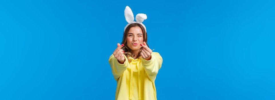 Holidays, traditions and celebration concept. Silly and cute happy blond girl having fun, showing korean heart gesture with fingers and send kisses, standing in rabbit ears, yellow hoodie.