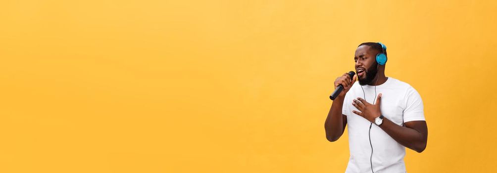 Portrait of cheerful positive chic. handsome african man holding microphone and having headphones on head listening music singing song enjoying weekend vacation isolated on yellow background.