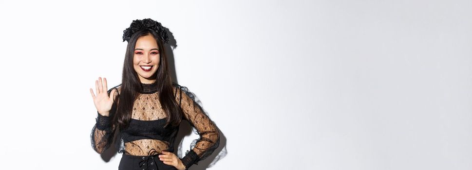 Image of friendly stylish asian woman in gothic lace dress waving hand to say hello, greeting someone, welcome people at halloween party, standing over white background.