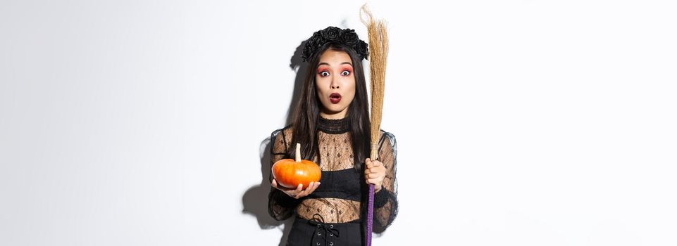 Image of surprised asian girl gasping wondered and stare at camera, wearing witch costume on halloween, holding broom and pumpkin, white background.