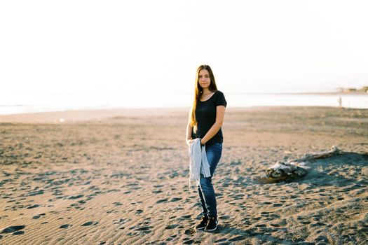 Girl in jeans with a sweater in her hands stands on a sandy beach by the sea. High quality photo