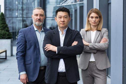 Successful and serious diverse team of three business people, man and woman focused looking at camera with arms crossed, portrait of co-workers outside office building
